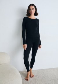Donna in piedi a piedi nudi su un tappeto, indossa una maglietta attillata a maniche lunghe nera e leggings, di fronte a una parete bianca semplice, con un divano beige nelle vicinanze.