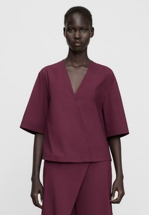 Femme noire portant un haut col en V de couleur vin avec des manches aux coudes et une jupe taille haute assortie sur un fond uni.