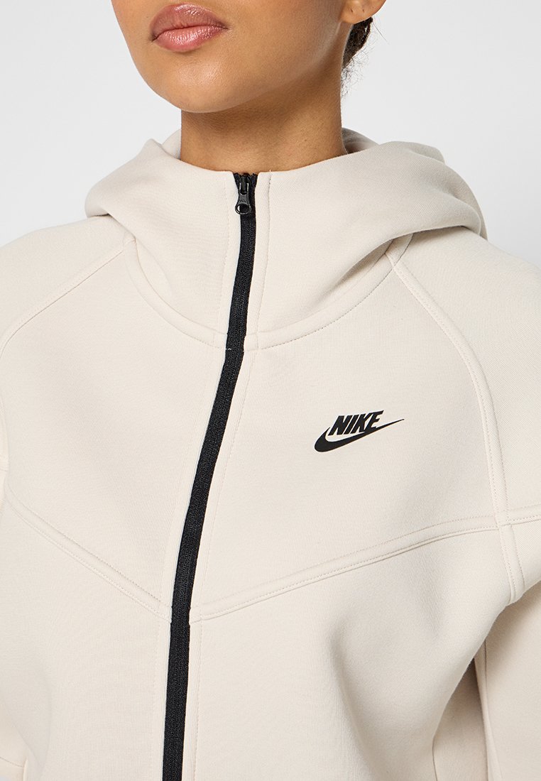 Felpa con zip beige dotata di cerniera nera e logo Nike. Presenta un cappuccio foderato, dettagli a coste e un tessuto liscio e strutturato.