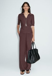 Top en tricot marron avec un col en V et des manches courtes, associé à un pantalon large assorti. Le modèle porte un grand sac fourre-tout en cuir noir.