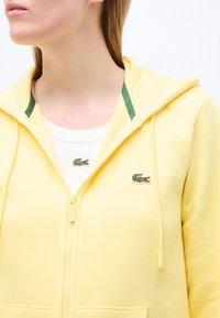 Femme portant un sweat à capuche jaune avec fermeture éclair sur un t-shirt blanc, tous deux arborant un petit logo de crocodile vert sur la poitrine.
