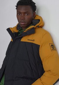 Timberland puffers jakke med en gul overdel og sort nederste sektion, med et logo-patch og fleece-foret hætte, designet til varme.