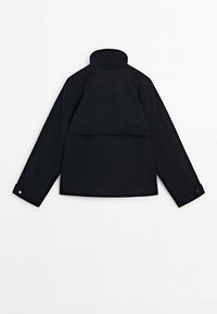 Veste noire à col avec manches longues, présentant un design à dos superposé et des poignets à boutons. Confectionnée dans un tissu lisse et résistant.