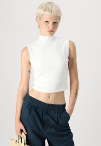 Top bianco corto senza maniche con collo alto, texture liscia e dettagli delle cuciture, abbinato a pantaloni blu scuro con vita elastica.
