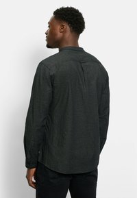 Mann, der nach links blickt, trägt ein dunkles langärmliges Hemd mit Knöpfen und schwarze Hosen vor einem schlichten weißen Hintergrund.