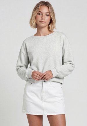 Tânără cu păr blond scurt, purtând un hanorac gri deschis și o fustă albă din denim, stând în fața unui fundal simplu.