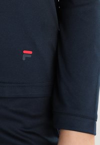 Maglietta sportiva blu navy con una texture liscia, caratterizzata da un piccolo logo rosso e grigio sulla manica e un design con polsino piegato.