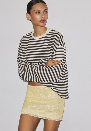 Kobieta z ciemnymi włosami zebranymi do tyłu, ubrana w czarno-białą, paskowaną bluzkę z długim rękawem oraz żółtą cekinową spódnicę z koronkową lamówką, z założonymi skrzyżowanymi ramionami.