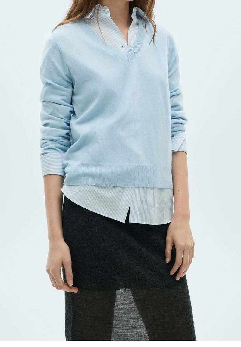 Femme portant un pull bleu clair à col en V par-dessus une chemise rayée claire, une jupe noire et des collants foncés, debout devant un fond uni.