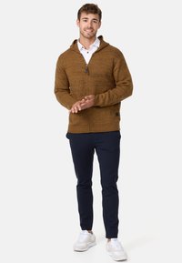 Brown zip-up hoodie with textured fabric, kangaroo pocket, and adjustable hood; paired with navy pants and white sneakers.