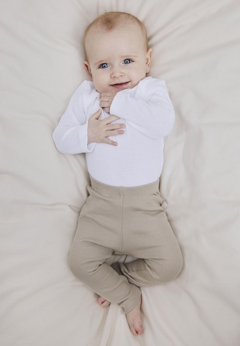 Hvit langermet body og beige rillete bukser på en baby som ligger på et lysfarget teppe, med en nøytral bakgrunn.