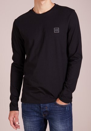 Mand iført sort langærmet trøje med lille "BOSS"-logo på brystet, kombineret med blå jeans, hånd i lommen.