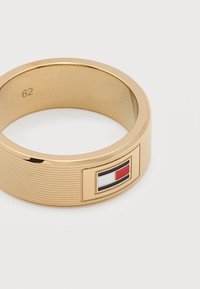 Δαχτυλίδι από μεταλλικό υλικό σε χρυσή απόχρωση με οριζόντιες ραβδώσεις και μικρό ορθογώνιο σήμα σημαίας σε κόκκινο, λευκό και μπλε στη εξωτερική πλευρά.