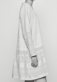 Biała sukienka z długimi rękawami, teksturowanymi wzorami i rozkloszowanym, falbaniastym dołem. Wykonana z lekkiego materiału w różnych wzorach pasków.