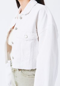 Valkoinen lyhyt farkkutakki, jossa on hopeiset napit, kaksi rintataskua ja hieman repsottava helma. Pehmeä pinta ja jämäkkä muotoilu.