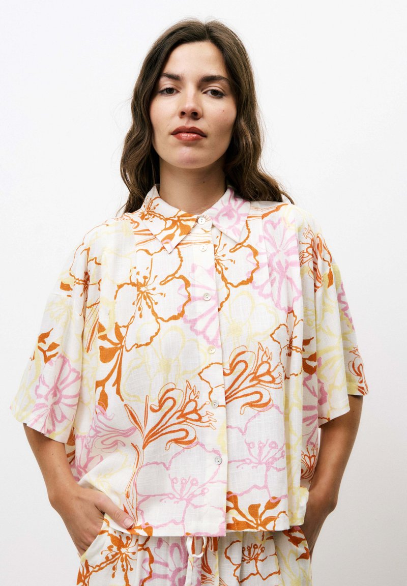 Korte mouw, knoopsluiting shirt in witte stof met bloemmotieven in oranje, roze en geel. Heeft een ontspannen pasvorm en een kraag.