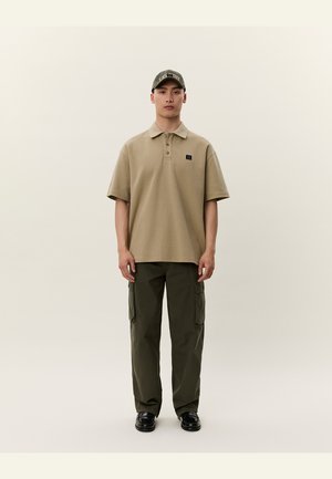 Polo de culoare bej cu guler și mâneci scurte, asortat cu pantaloni cargo de culoare verde măsliniu, cu buzunare laterale, și pantofi negri.