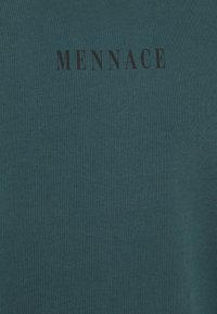 T-shirt in cotone teal con texture a coste, caratterizzato dalla parola "MENNACE" in lettere maiuscole nere sul petto.