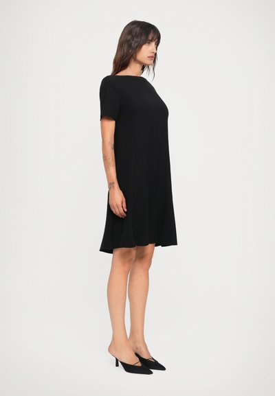Black short-sleeve dress with a loose fit and round neckline, made from smooth fabric. Worn with pointed black heels, showcasing a relaxed silhouette.
