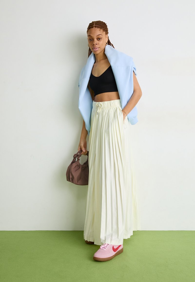 Giacca oversized blu chiaro sopra un crop top nero, abbinata a una gonna maxi plissettata crema e sneakers rosa. Borsa a tracolla marrone con cordoncino.