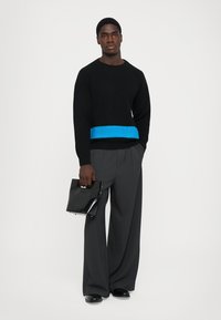 Mann trägt einen schwarzen Pullover mit blauer Streifen, weite graue Hose, schwarze Schuhe und hält eine glänzende schwarze Handtasche vor einem weißen Hintergrund.