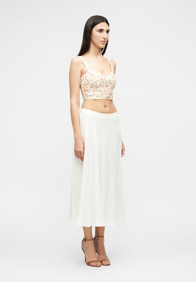 Γυναίκα με μακριά σκούρα μαλλιά που φοράει φλοράλ τοπ, λευκή midi φούστα με ψηλή μέση και ταμπά πέδιλα με λουράκι στον αστράγαλο, στέκεται μπροστά από απλό φόντο.