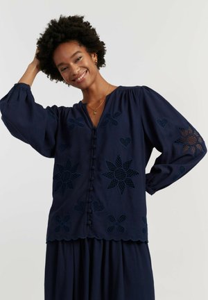 Femme souriante aux cheveux bouclés portant un chemisier bleu marine avec boutons sur le devant, ourlet festonné et motifs découpés floraux et en forme de cœur, assorti à une jupe.