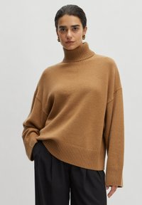 Brauner Rollkragenpullover mit lockerer Passform und geripptem Saum. Der Stoff wirkt weich und strukturiert und wird mit schwarzen Hosen kombiniert.