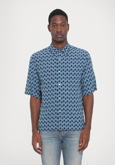 Jonge man die rechtop staat en een blauw patroon met korte mouwen overhemd en lichtblauwe jeans draagt, tegen een effen achtergrond.