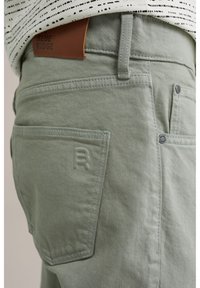 Pantaloni din bumbac de culoare verde deschis, cu o etichetă din piele inscripționată cu "BLUE RIDGE." Caracteristicile includ detalii la buzunarul din spate și o textură subtilă.