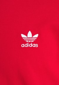 Červená látka s hladkým texturem, se středem bílého vyšívaného loga Adidas a designem se třemi listy.