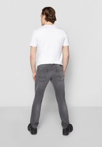 Bărbat în picioare, cu spatele la noi, purtând un tricou alb, blugi gri și pantofi negri, pe un fundal simplu, deschis.