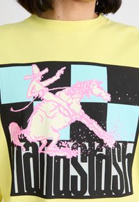 Sweatshirt en coton jaune avec un grand graphisme d'un cow-boy sur un cheval en rose, sur un fond à damier noir et bleu clair.