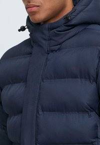 Marineblå pufferjakke med høy krage, med synlige quiltede seksjoner, snordtrekkjusteringer på hetten, og jevn utvendig tekstur.