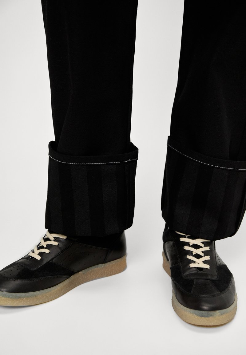 Schwarze Hosen mit gestreiften Bündchen und weißen Nähten, kombiniert mit schwarzen Sneakers mit einer Gummisohle und beigen Schnürsenkeln.