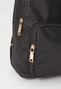 Zaino in nylon nero con hardware della cerniera dorato, dotato di due cerniere sulla tasca frontale e materiale liscio e testurizzato.