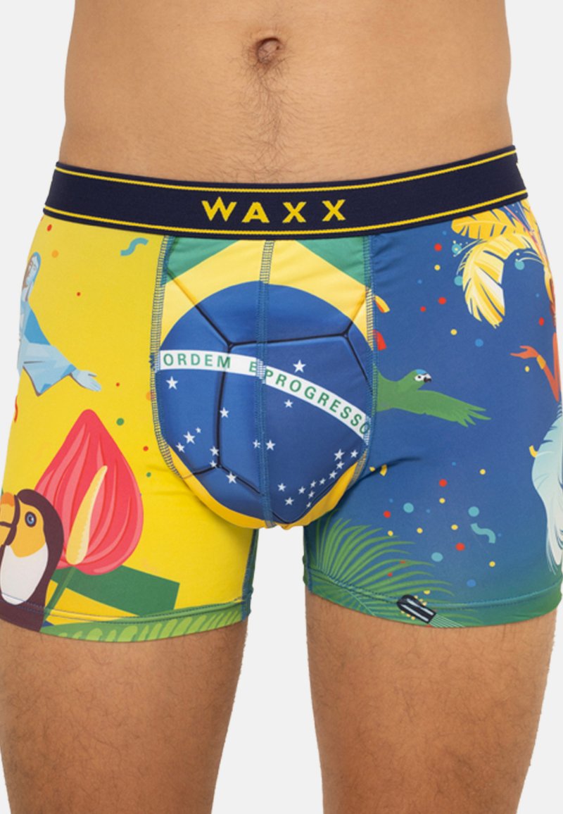 Boxer shorts colorés avec un design tropical, une base jaune, des accents bleus et des illustrations d'animaux et de fleurs. La ceinture porte l'inscription "WAXX."