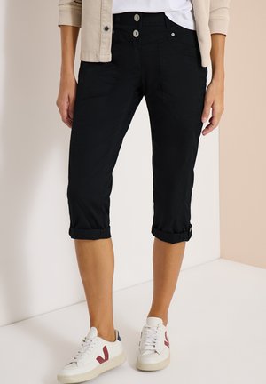 Femme portant un pantalon capri noir avec revers et fermeture double bouton, veste beige, baskets blanches avec logo rouge en forme de "V", debout à l'intérieur.