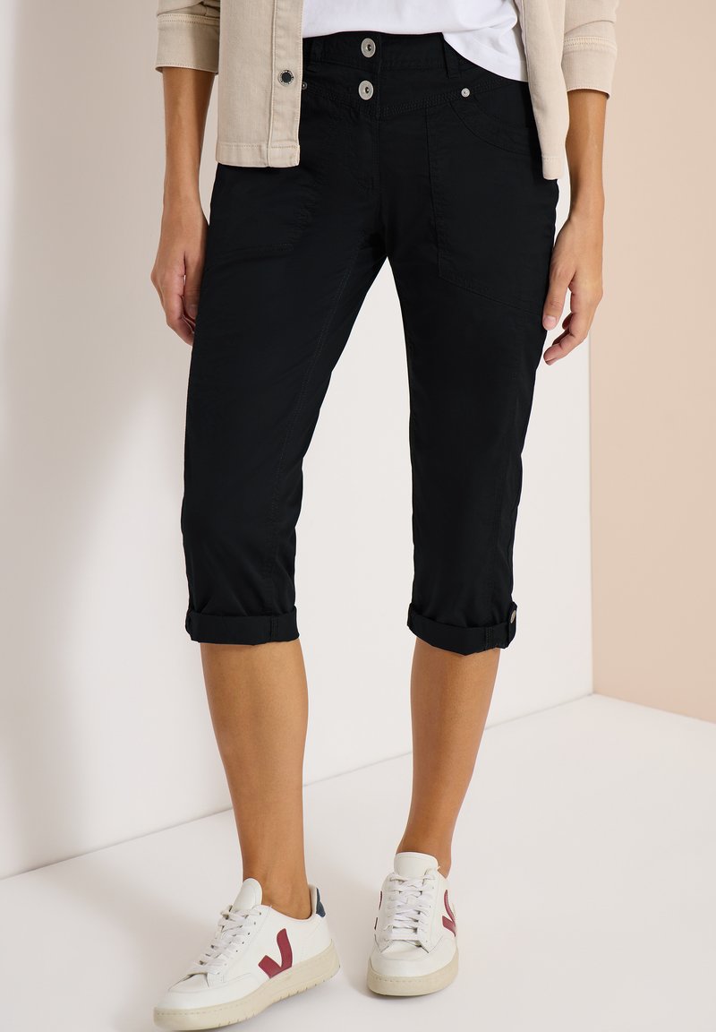Femme portant un pantalon capri noir avec revers et fermeture double bouton, veste beige, baskets blanches avec logo rouge en forme de "V", debout à l'intérieur.
