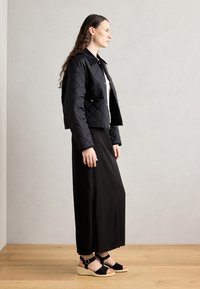 Veste courte noire matelassée avec un col, associée à une longue robe noire et des sandales compensées noires. Design minimaliste et détails de texture.