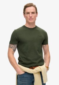 T-shirt in cotone verde oliva con maniche corte, collo a giro e un piccolo dettaglio con logo. Indossato con jeans blu e un maglione a maglia di colore chiaro legato in vita.
