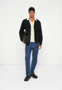 Zwarte gebreide cardigan over een beige opstaande kraag poloshirt, gecombineerd met blauwe jeans en zwarte schoenen, met een zwarte tas in de hand. Effen witte achtergrond.