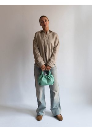 Mujer con camisa beige abotonada y vaqueros de pierna ancha azul claro, sosteniendo un bolso pequeño verde recogido frente a un fondo liso.