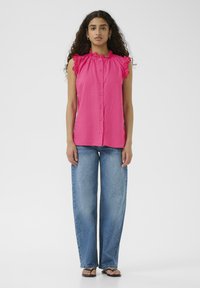 Top senza maniche rosa con dettagli arricciati sulle spalle, chiusura con bottoni, abbinato a jeans in denim blu a gamba larga e sandali neri piatti.