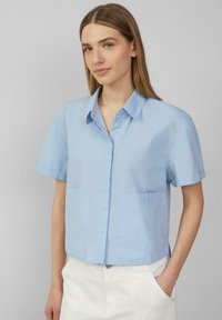 Camicetta azzurra chiara a maniche corte con colletto, due tasche frontali e una trama liscia. Fit casual e ampio.