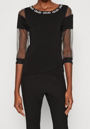 Femme portant un haut noir avec des manches trois-quarts transparentes et scintillantes et "LIU JO" imprimé autour du col et des poignets, associé à un pantalon noir.