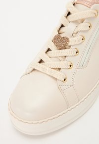 Ljust beige lädersneaker med en glansig roséguldfinish, med krämfärgade snören, guldfärgade öglor och en dekorativ hjärtformad nitar på snörena.