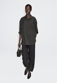 Dunkles, übergroßes Hemd kombiniert mit lockeren schwarzen Hosen. Accessoires sind eine kleine, geflochtene schwarze Handtasche und spitze schwarze Schuhe.