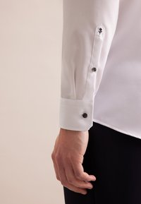 Fekete gombbal ellátott, karcsú fazonú fehér ing, virágmintás hímzéssel a ruha ujjának szellőzője közelében.