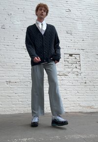 Black cable-knit cardigan over a white shirt, gray wide-leg pants, and gray-black sneakers with teal accents, standing against a white brick wall.